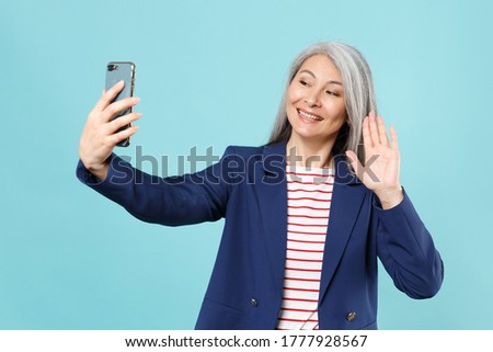 Smiling gray-haired business woman in blue suit isolated on blue background. Achievement career wealth business concept. Doing selfie shot on mobile phone making video call, waving greeting with hand