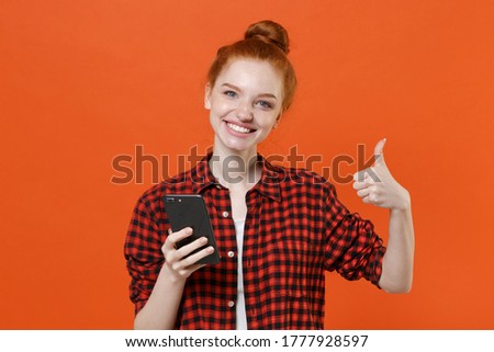 Smiling young readhead girl in casual red checkered shirt posing isolated on orange wall background studio portrait. People lifestyle concept. Mock up copy space. Using mobile phone, showing thumb up