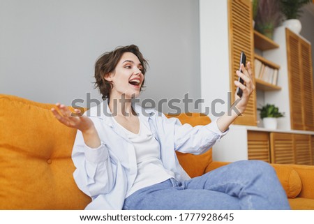 Excited young woman girl in casual clothes sitting on couch spending time in living room at home. Rest relax good mood leisure lifestyle concept. Doing selfie shot on mobile phone, making video call