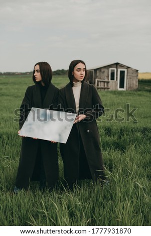 Portrait of beautiful young brunette women sisters with long hair in a coat and jeans in a green field with a mirror. Stylish fashion photography, clothing advertising.