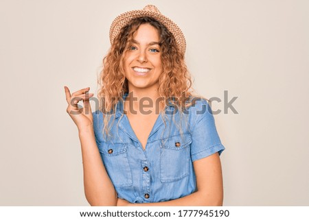 Young beautiful woman with blue eyes wearing casual denim dress and summer hat with a big smile on face, pointing with hand and finger to the side looking at the camera.