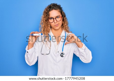 Young beautiful blonde doctor woman with blue eyes wearing stethoscope holding thermometer with angry face, negative sign showing dislike with thumbs down, rejection concept