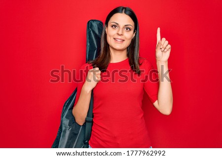 Young beautiful brunette guitarist woman holding guitar case over isolated red background surprised with an idea or question pointing finger with happy face, number one