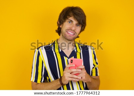 Pleased Caucasian man standing over yellow background, wearing casual clothes using self phone and looking and winking at the camera. Flirt and coquettish concept.