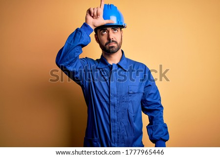 Mechanic man with beard wearing blue uniform and safety helmet over yellow background making fun of people with fingers on forehead doing loser gesture mocking and insulting.