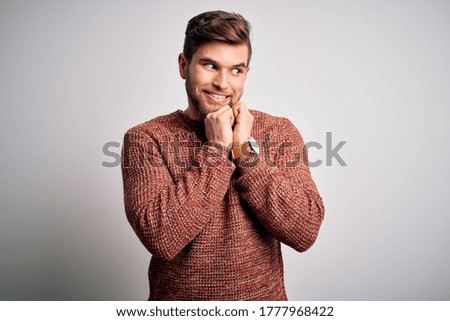 Young blond man with beard and blue eyes wearing casual sweater over white background laughing nervous and excited with hands on chin looking to the side