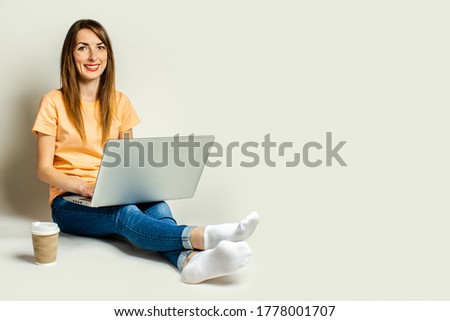 Smiling young woman holding a laptop on her knees, a paper cup with coffee is standing nearby on a light background