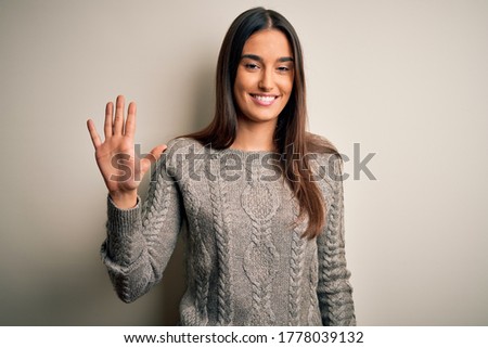 Young beautiful brunette woman wearing casual sweater over isolated white background showing and pointing up with fingers number five while smiling confident and happy.