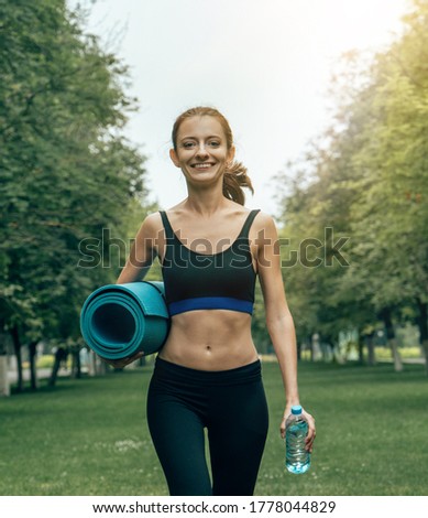 outdoor portrait of beautiful young smiling caucasian sportswoman girl walking in the park with a fitness mat in her hands and a bottle of water on a summer sunny day. healthy lifestyle concept