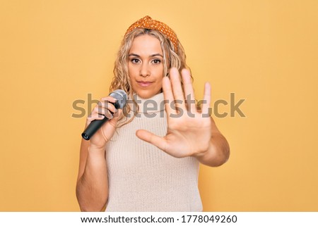 Young beautiful blonde singer woman singing using microphone over yellow background with open hand doing stop sign with serious and confident expression, defense gesture
