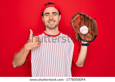 Young handsome sporty man with blue eyes playing baseball using glove and ball happy with big smile doing ok sign, thumb up with fingers, excellent sign