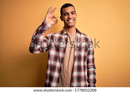 Young handsome african american man wearing casual shirt standing over yellow background smiling positive doing ok sign with hand and fingers. Successful expression.