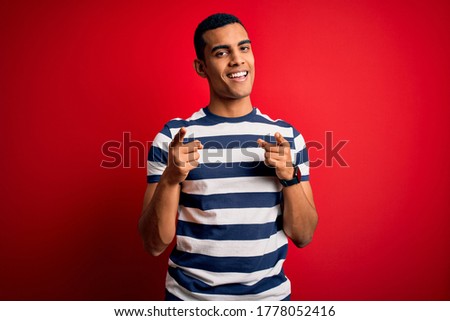 Handsome african american man wearing casual striped t-shirt standing over red background pointing fingers to camera with happy and funny face. Good energy and vibes.