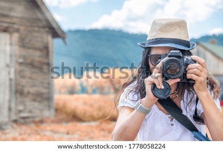 Woman wearing straw hat making pictures of mountain landscpae with wooden hut.