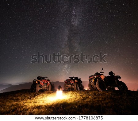 Night picture. Three atv quad motorbikes standing on the top of mountain near burning campfire, under beautiful night sky full of stars and Milky way on background