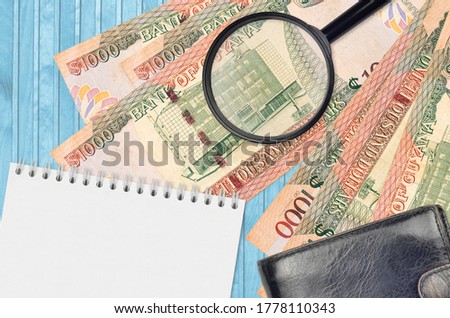 1000 Guyanese dollars bills and magnifying glass with black purse and notepad. Concept of counterfeit money. Search for differences in details on money bills to detect fake