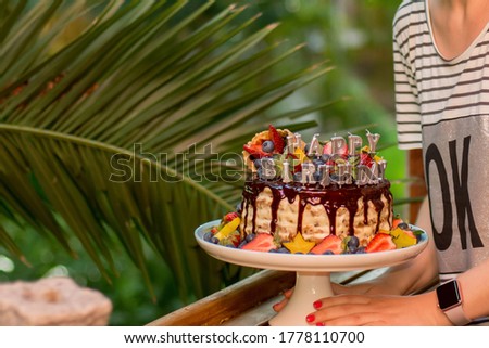 Selective focus image of hands holding layered cake decorated with sparkles, happy birthday candles, chocolate glaze, strawberries, blueberries, carambola star fruits on tropical background