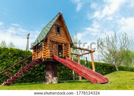 Small wood log playhouse hut with stairs ladder and wooden slide on children playground at park or house yard. Green grass lawn garden and blue clear sky in background on bright sunny summer day Royalty-Free Stock Photo #1778124326