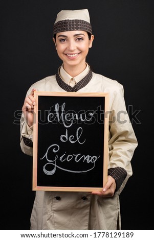 Smiling chef in uniform holds a blackboard in his hand with on it written in Italian "Menu del giorno" which means "Menu of the day", isolated on orange background
