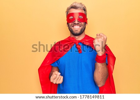 Handsome blond man wearing super hero costume with mask and cape over yellow background celebrating surprised and amazed for success with arms raised and eyes closed
