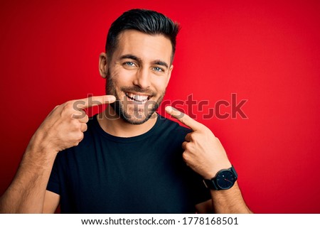 Young handsome man wearing casual black t-shirt standing over isolated red background smiling cheerful showing and pointing with fingers teeth and mouth. Dental health concept.