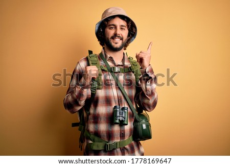 Young hiker man with curly hair and beard hiking wearing backpack and water canteen with a big smile on face, pointing with hand finger to the side looking at the camera.