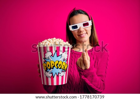Young beautiful girl watching movie using 3d glasses eating box with popcorns doing money gesture with hands, asking for salary payment, millionaire business
