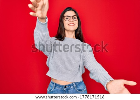 Young beautiful brunette woman wearing casual sweater over red background looking at the camera smiling with open arms for hug. Cheerful expression embracing happiness.
