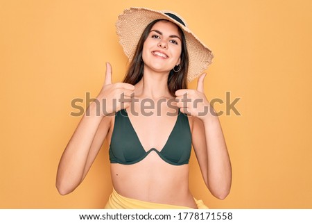 Young beautiful girl wearing swimwear bikini and summer sun hat over yellow background approving doing positive gesture with hand, thumbs up smiling and happy for success. Winner gesture.