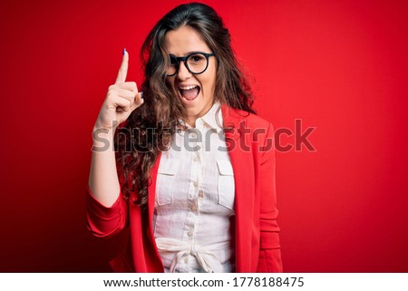 Young beautiful woman with curly hair wearing jacket and glasses over red background pointing finger up with successful idea. Exited and happy. Number one.