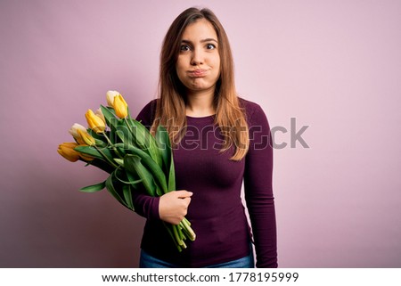 Young blonde woman holding romantic bouquet of yellow tulips flowers over pink background puffing cheeks with funny face. Mouth inflated with air, crazy expression.