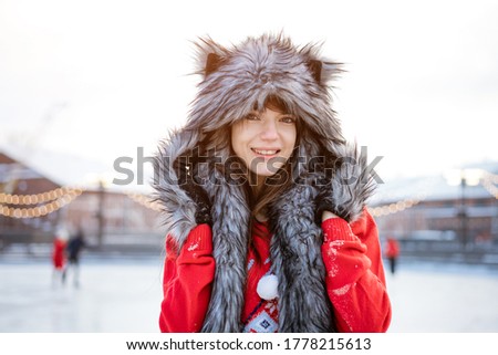 Happy young woman in a wolf hat in winter at the ice rink poses in a red sweater outside in the afternoon