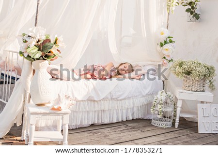 Delicate little blonde girl with long hair in a beautiful pink powdery dress sleeps in a white bed with chiffon canopies, decorated with wisteria, flowers against the background of the sea. Magic day