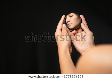 Hispanic woman looking at herself in a mirror with black background - close up hands holding a small mirror with the reflection of a face in it - reflection of her face