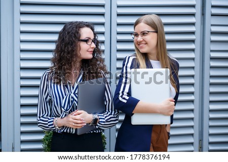 Portrait of two women businessmen who are standing with laptops in their hands, new project. Young business colleagues discuss office work. Successful female manager with glasses. Business concept