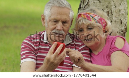 Family weekend picnic. Old people, merry senior caucasian couple using smartphone. Online shopping, browsing on phone. Social media network. Elderly man, woman. Chatting, social media network