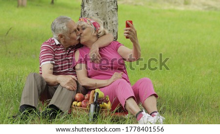 Family weekend picnic. Old people, merry senior caucasian couple using smartphone makes selfie. Remote online video chat call on phone. Elderly man, woman. Husband and wife in park. Grandparents