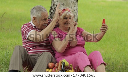 Family weekend picnic. Old people. Senior caucasian couple using smartphone for video call. Talking on webcam. Chatting, social media network on phone. Elderly man, woman. Husband and wife in park