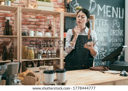Portrait of young barista woman standing behind counter thoughtfully looking on wooden table at cafe bar. pensive female waitress in apron and white shirt in coffee shop holding note frowning face.