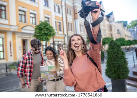 Taking picture, mood. Happy smiling girl with open mouth gesturing taking selfie on camera, friends at a distance