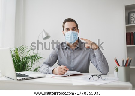 Excited young business man in gray shirt sit at desk work on laptop in light office on white wall background. Achievement business career concept. Writing notes in notebook point on sterile face mask