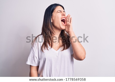 Young beautiful brunette woman wearing casual t-shirt standing over isolated white background shouting and screaming loud to side with hand on mouth. Communication concept.