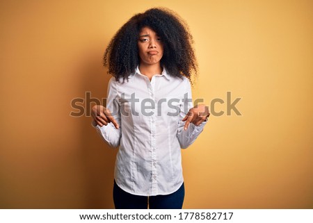 Young beautiful african american elegant woman with afro hair standing over yellow background Pointing down looking sad and upset, indicating direction with fingers, unhappy and depressed.