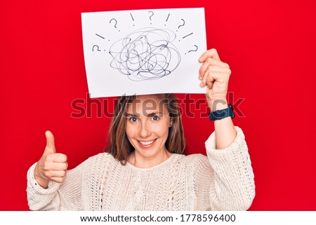 Young beautiful blonde woman holding picture of chaos above head smiling happy and positive, thumb up doing excellent and approval sign 