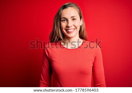 Young beautiful blonde woman wearing casual t-shirt standing over isolated red background with a happy and cool smile on face. Lucky person.