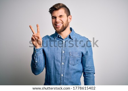 Young handsome blond man with beard and blue eyes wearing casual denim shirt smiling with happy face winking at the camera doing victory sign with fingers. Number two.