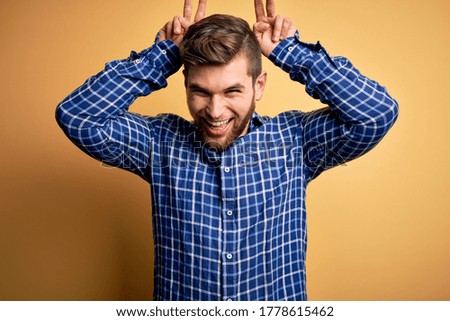 Young blond businessman with beard and blue eyes wearing shirt over yellow background Posing funny and crazy with fingers on head as bunny ears, smiling cheerful