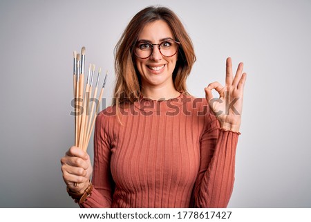 Young beautiful brunette artist woman holding painter brushes over white background doing ok sign with fingers, excellent symbol