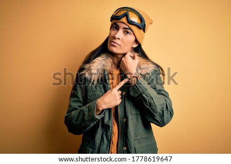 Young beautiful hispanic woman wearing ski glasses and coat for winter weather In hurry pointing to watch time, impatience, looking at the camera with relaxed expression