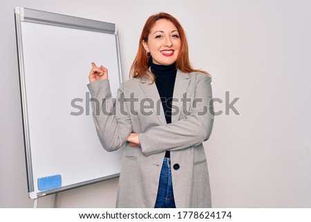 Young beautiful redhead businesswoman doing business presentation using magnetic board with a big smile on face, pointing with hand and finger to the side looking at the camera.
